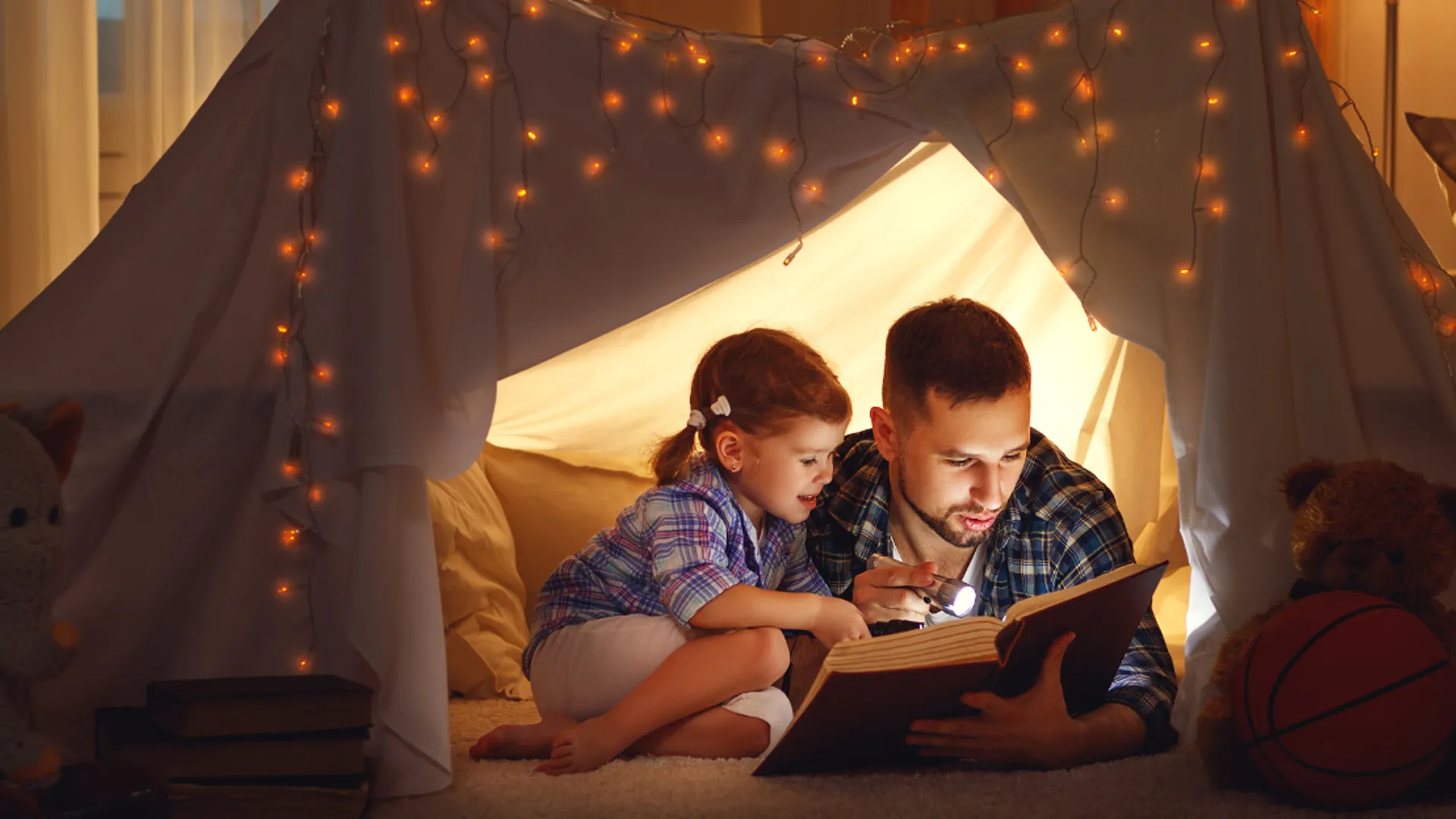 Bild på dotter och pappa som ligger och läser bok i ett tält upplyst tält.