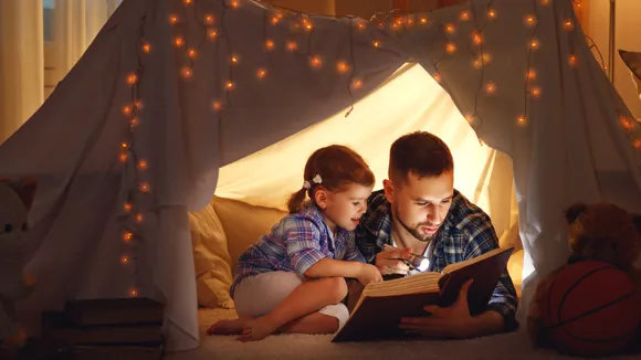 Pappa och dotter läser bok i ett upplyst tält inomhus.