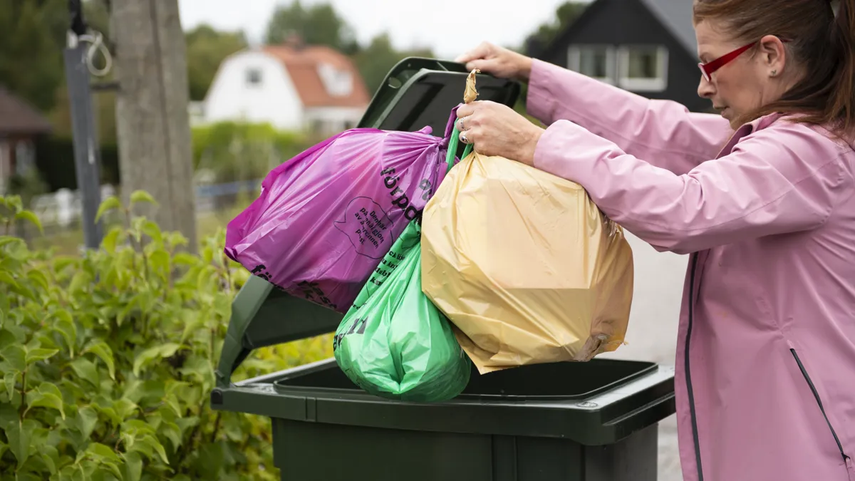En kvinna i rosa jacka slänger tre olikfärgade påsar i en soptunna. 