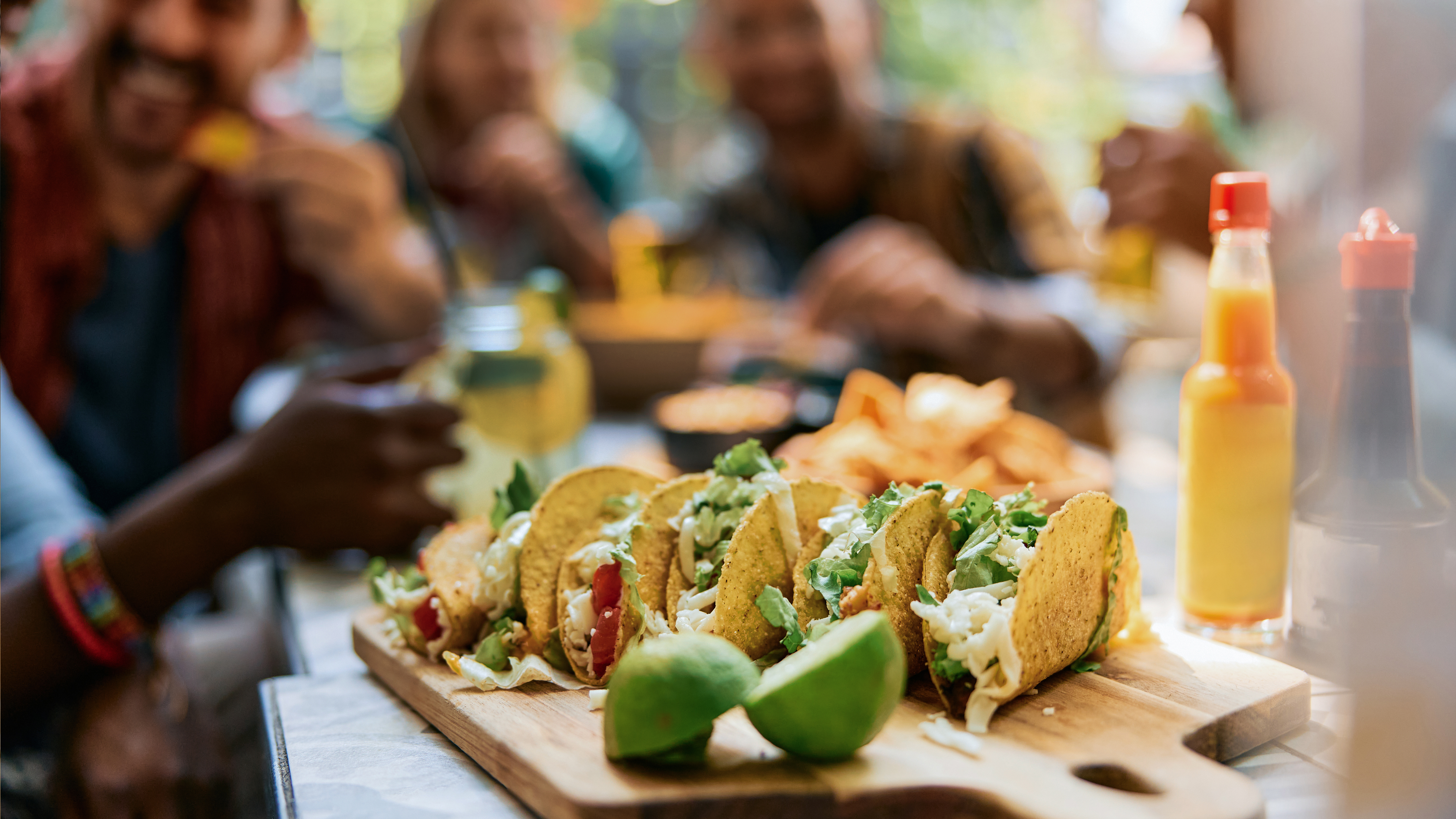 Flera färdiga tacos på en bricka tillsammans med lime med människor i bakgrunden som har trevlig tillsammans.