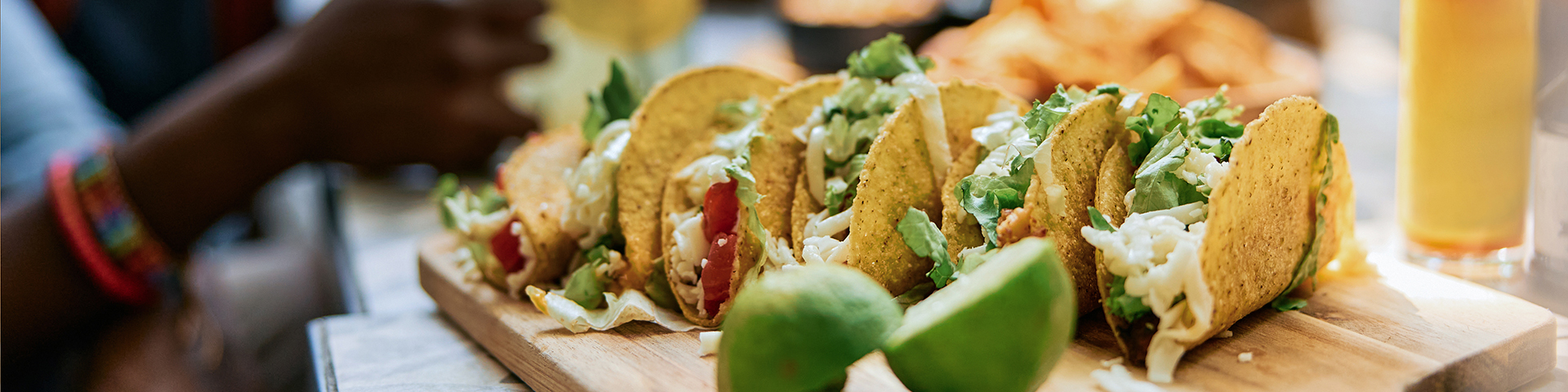 Flera färdiga tacos på en bricka tillsammans med lime, med människor i bakgrunden som har trevlig tillsammans.