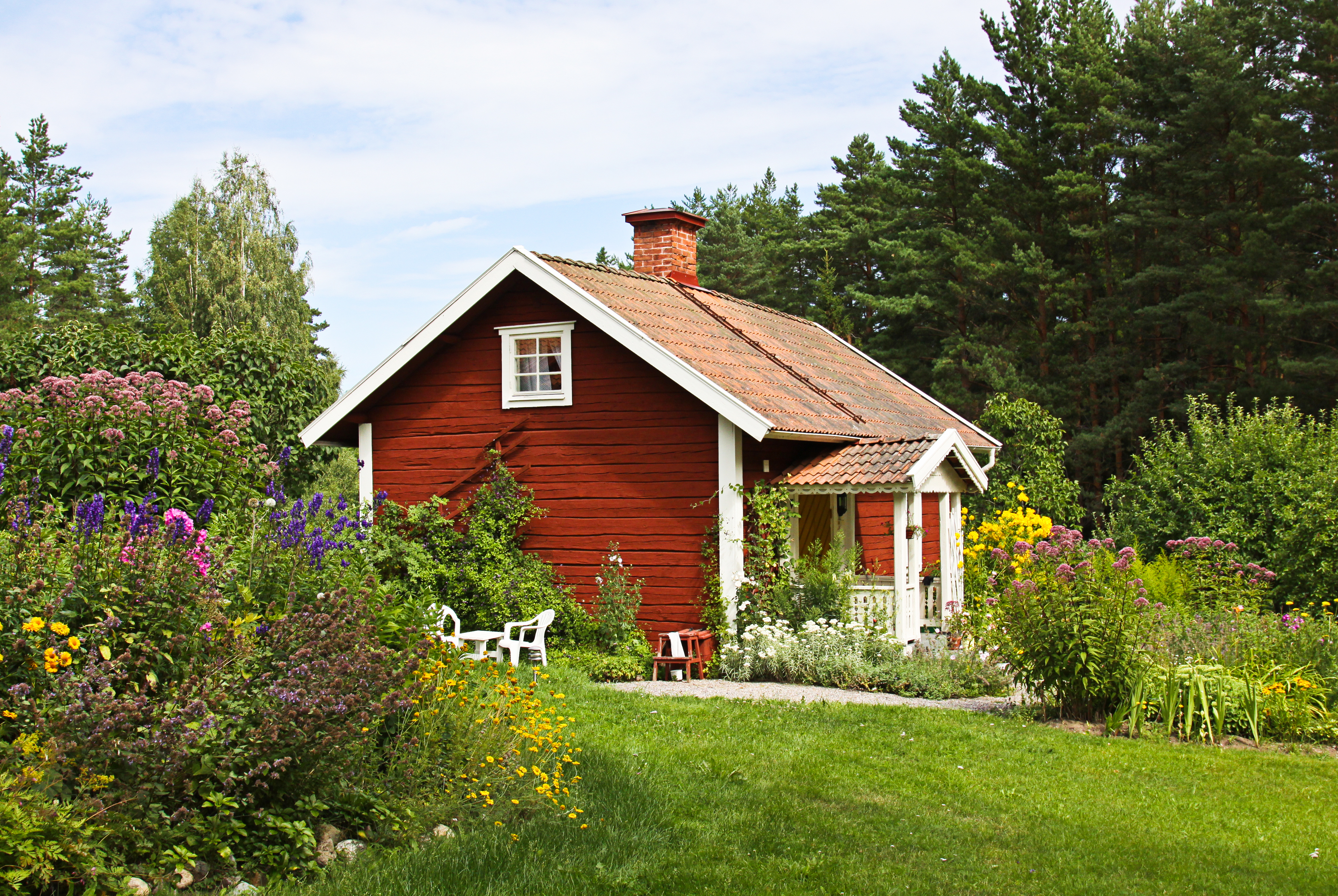 Rött hus med veranda i grön trädgård, omgiven av blomster och träd.