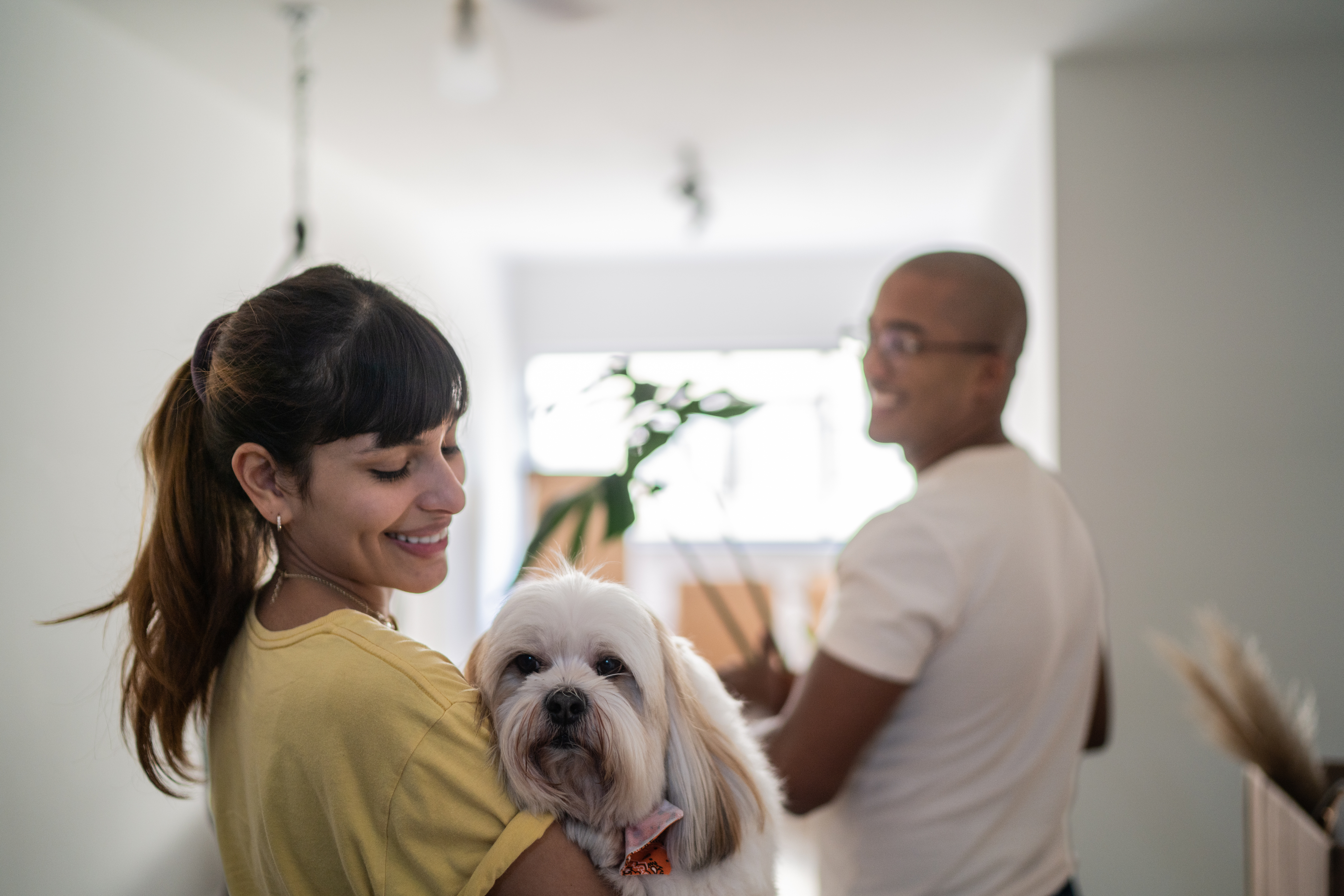 Leende kvinna inomhus med hund i famnen, i bakgrunden skymtar en leende man.