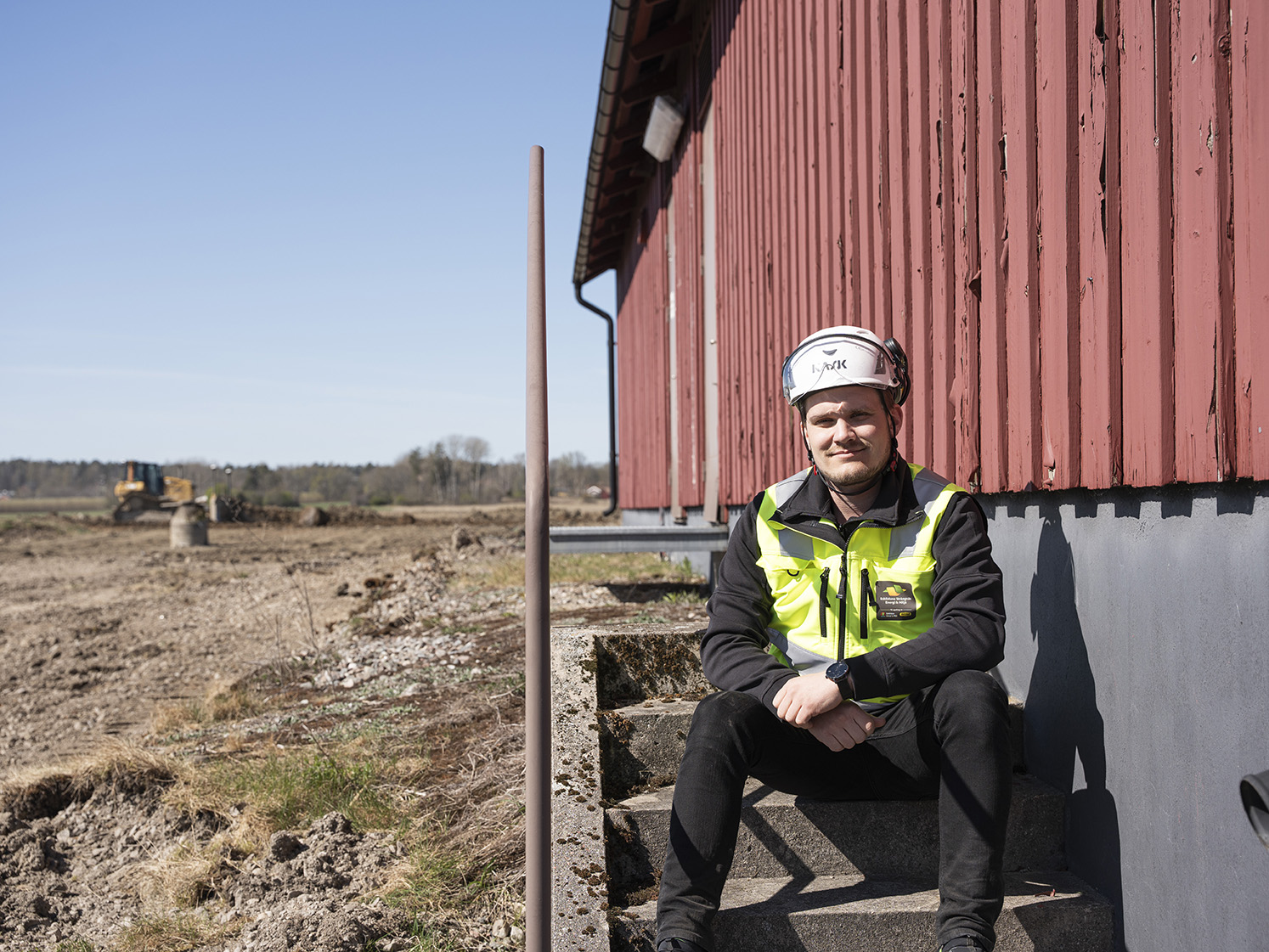 Projektledaren Jimmay sitter på en trappa iklädd varselväst och hjälm. I bakgrunden på ett fält syns en grävmaskin.