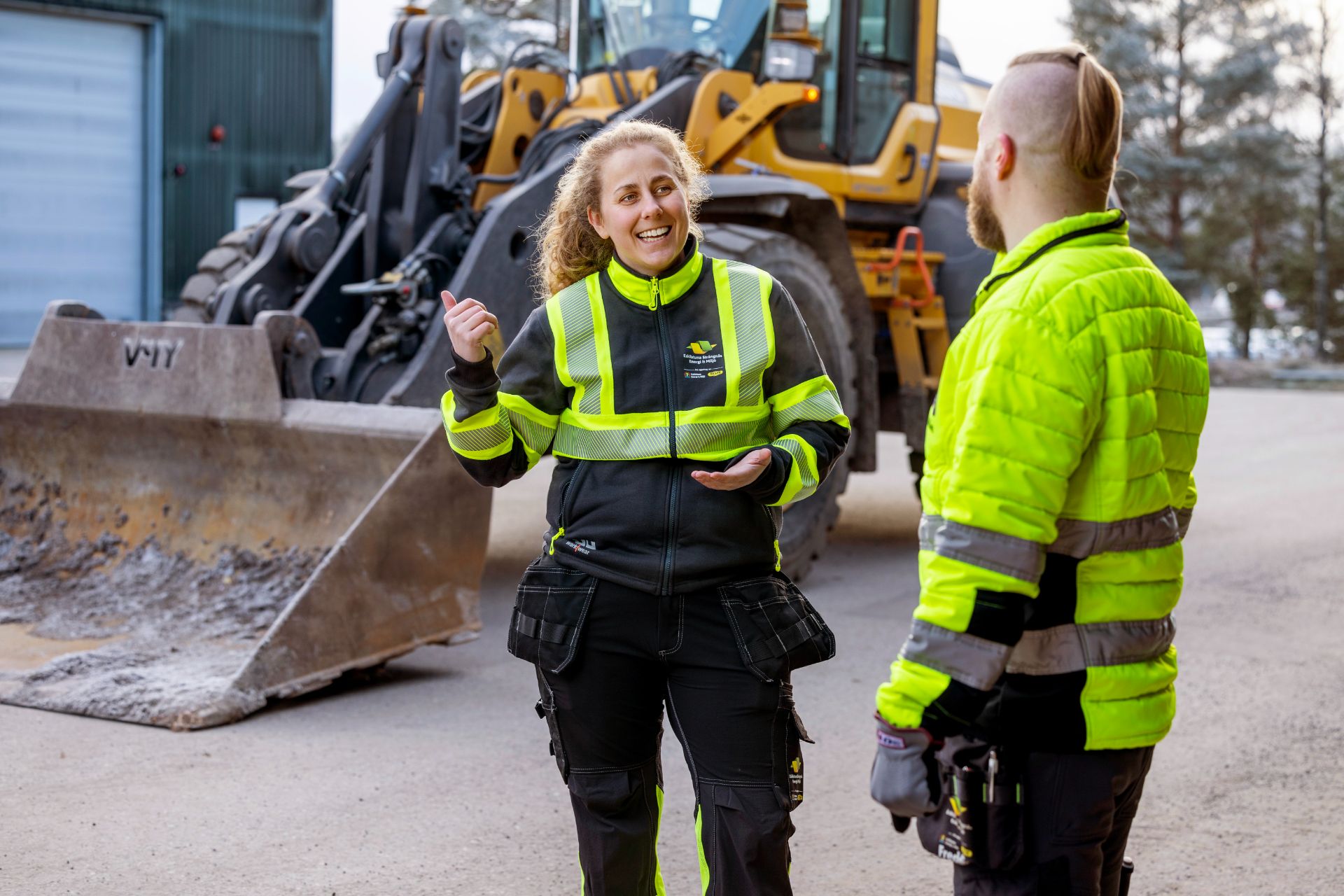 Två personer pratar med varandra vid en grävmaskin.