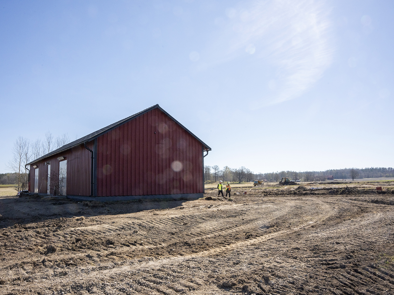 Röd byggnad på en jord- och sandblandad plan. Två personer i skyddskläder och ett par maskiner syns på långt håll.